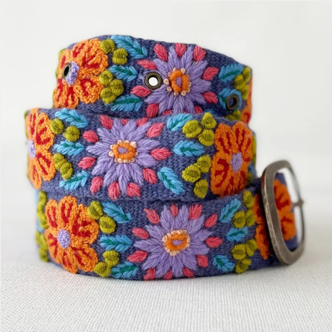 The JENNY KRAUSS - PRAIRIE FLOWERS EMBROIDERED WOOL BELT features colorful floral embroidery in orange, purple, green, and blue on wool, handcrafted in Peru with a silver buckle, shown rolled up on a light surface.