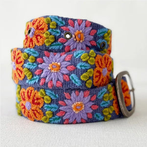 The JENNY KRAUSS - PRAIRIE FLOWERS EMBROIDERED WOOL BELT features colorful floral embroidery in orange, purple, green, and blue on wool, handcrafted in Peru with a silver buckle, shown rolled up on a light surface.