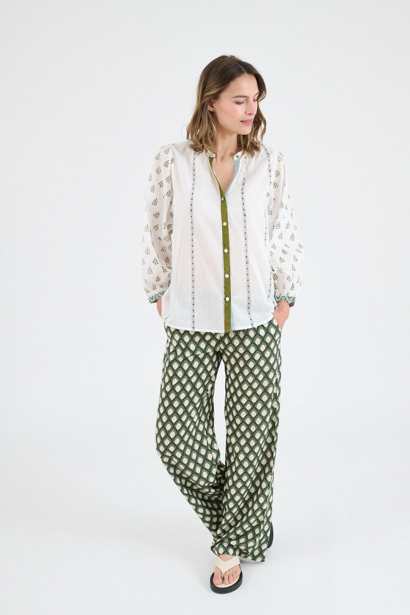 A woman wears the STORIATIPIC LUCIE FLOWER BLOUSE—a cotton blouse with patterned sleeves and green trim—paired with wide-leg green patterned pants, standing against a white background in sandals, evoking casual elegance.