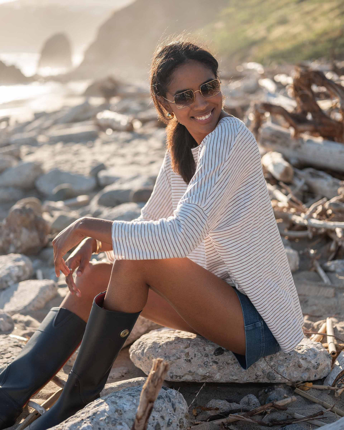 A woman with wavy brown hair models the MERSEA - CATALINA SLUB TEE—an oversized, long-sleeved white top with thin gray stripes—by MERSEA. She pairs it with blue jeans and stands against a plain, light background, hands behind her back.