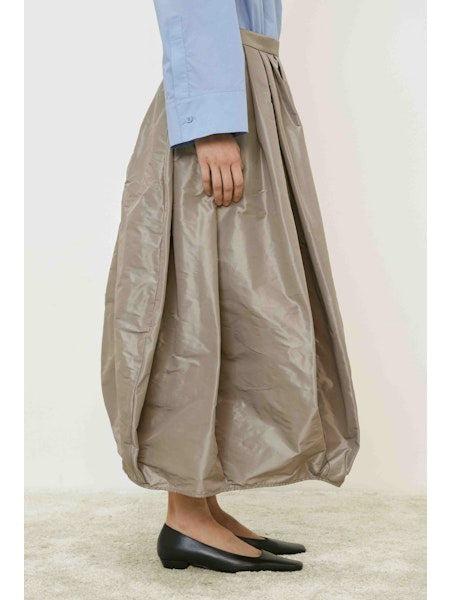 A person wears the MEIMEIJ Taffeta Balloon Skirt in shiny taupe, paired with a light blue long-sleeve shirt and black flats, standing on a light carpet against a white background.