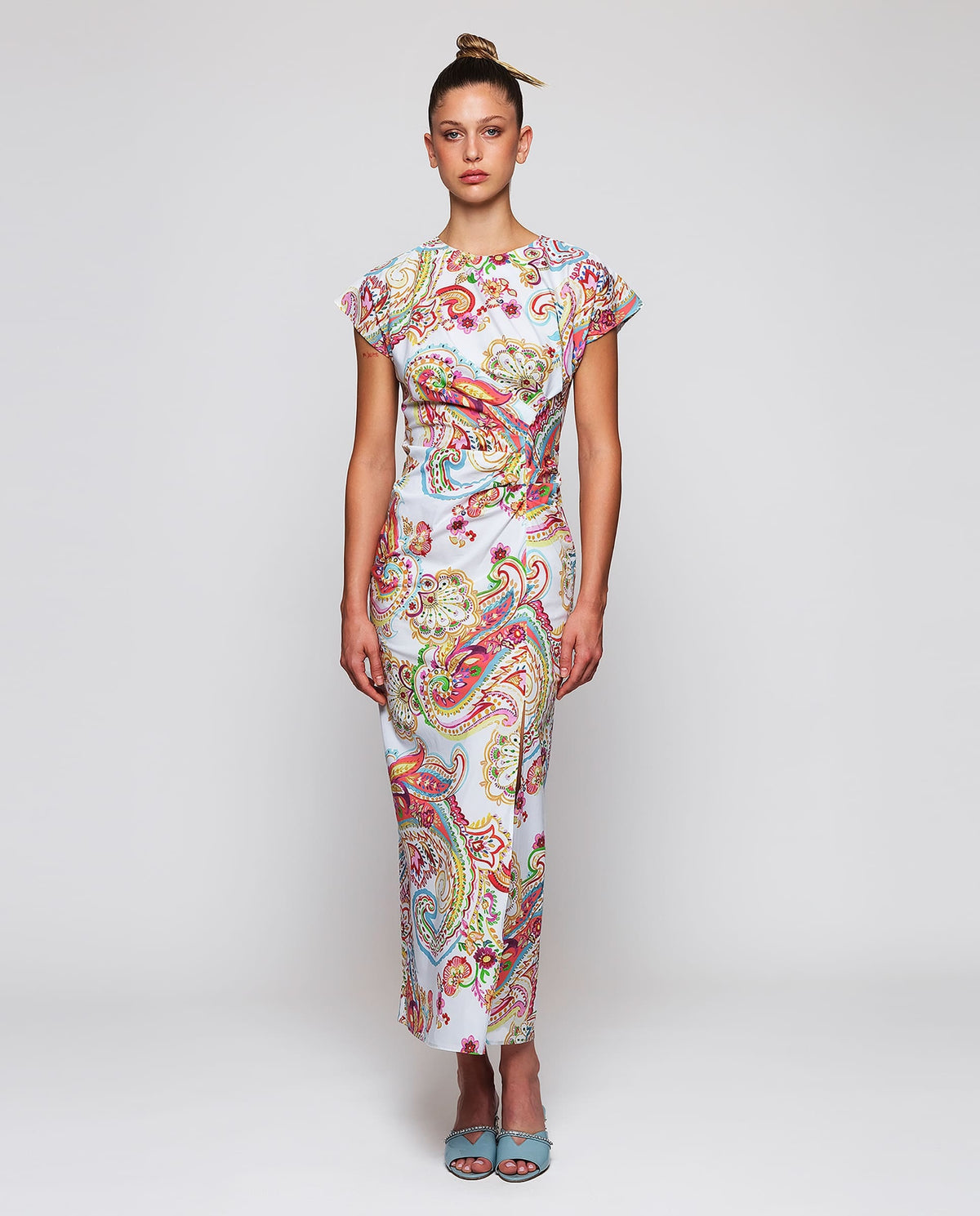 A woman stands with her back to the camera in a MIRTO 1956 PAISLEY PRINTED MIDI DRESS, featuring short sleeves, paired with light blue low-heeled shoes. Her hair is styled in a neat bun, set against a plain white background.