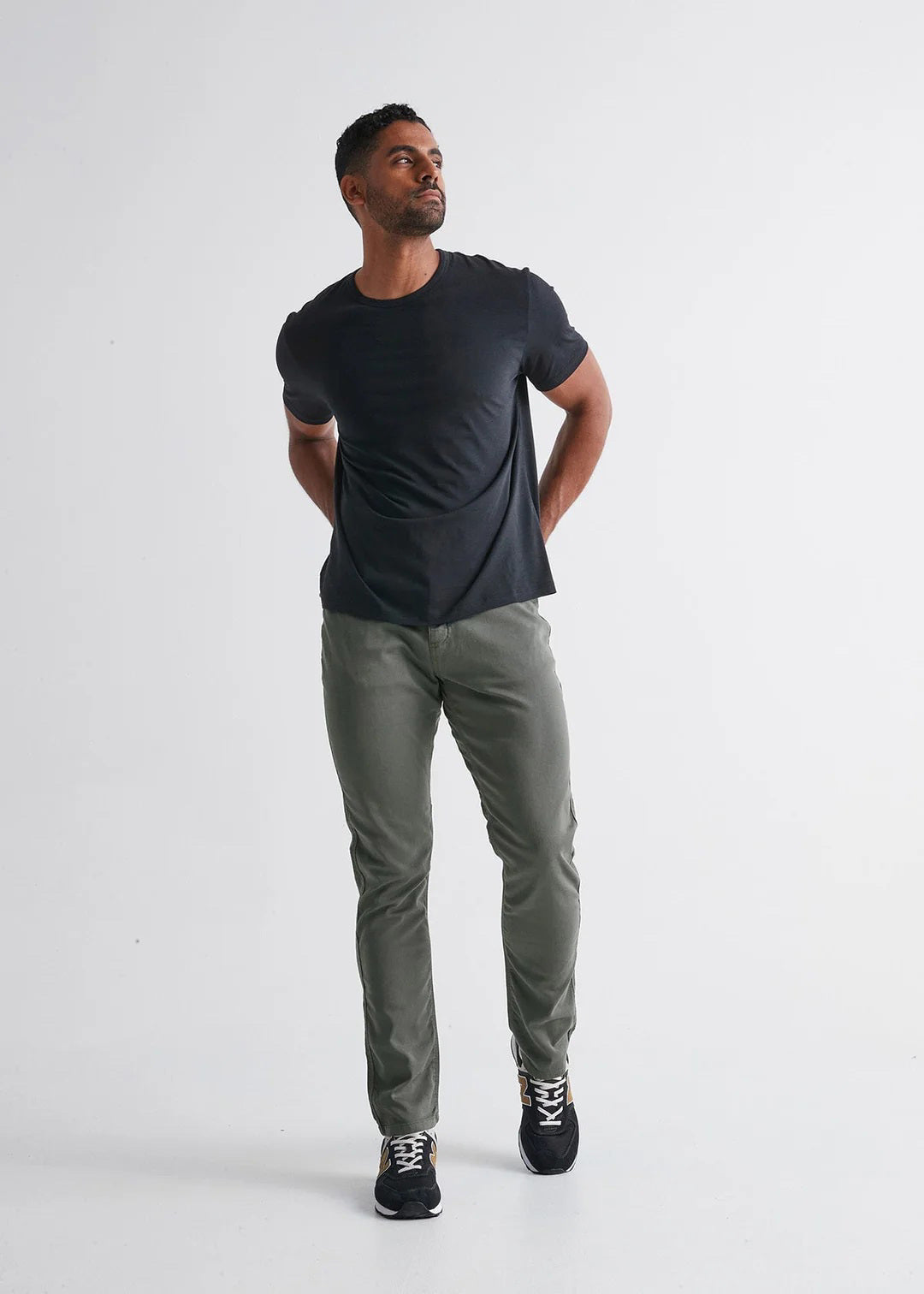 A person stands against a plain white background wearing DUER No Sweat Relaxed Taper Pants in Gull (olive green), a black T-shirt, and black New Balance sneakers with white soles and gold accents.