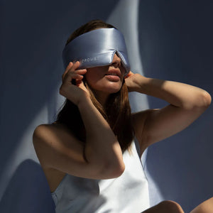 An older person with long gray hair wears a luxurious beige DROWSY - SATIN SLEEP MASK by DROWSY SLEEP CO, made of mulberry silk. In a matching satin top, they stand against a plain white background, ready for beauty sleep.
