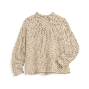 A woman in a sunlit kitchen smiles, wearing the FRANK & EILEEN Monterey Pointelle Rolled Funnel Neck Cotton Sweater in Sand with blue jeans; its machine washable design brings comfort and ease to her day.