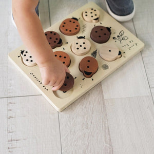 The WEE GALLERY - LADYBUGS COUNT TO 10 TRAY PUZZLE by WEE GALLERY is a wooden puzzle board featuring ladybug-shaped pieces in varying shades from light beige to dark brown. This educational puzzle helps teach counting skills and depicts leaves and twigs alongside the words "Count to Ten" on the left side. Each ladybug is adorned with black dots on their backs to represent number concepts.