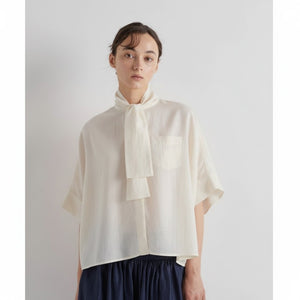 Someone stands against a plain light background wearing the MEIMEIJ SCARF TIE NECK BLOUSE—an oversized cream top with a tie-neck, chest pocket, and short sleeves—paired with a dark skirt.