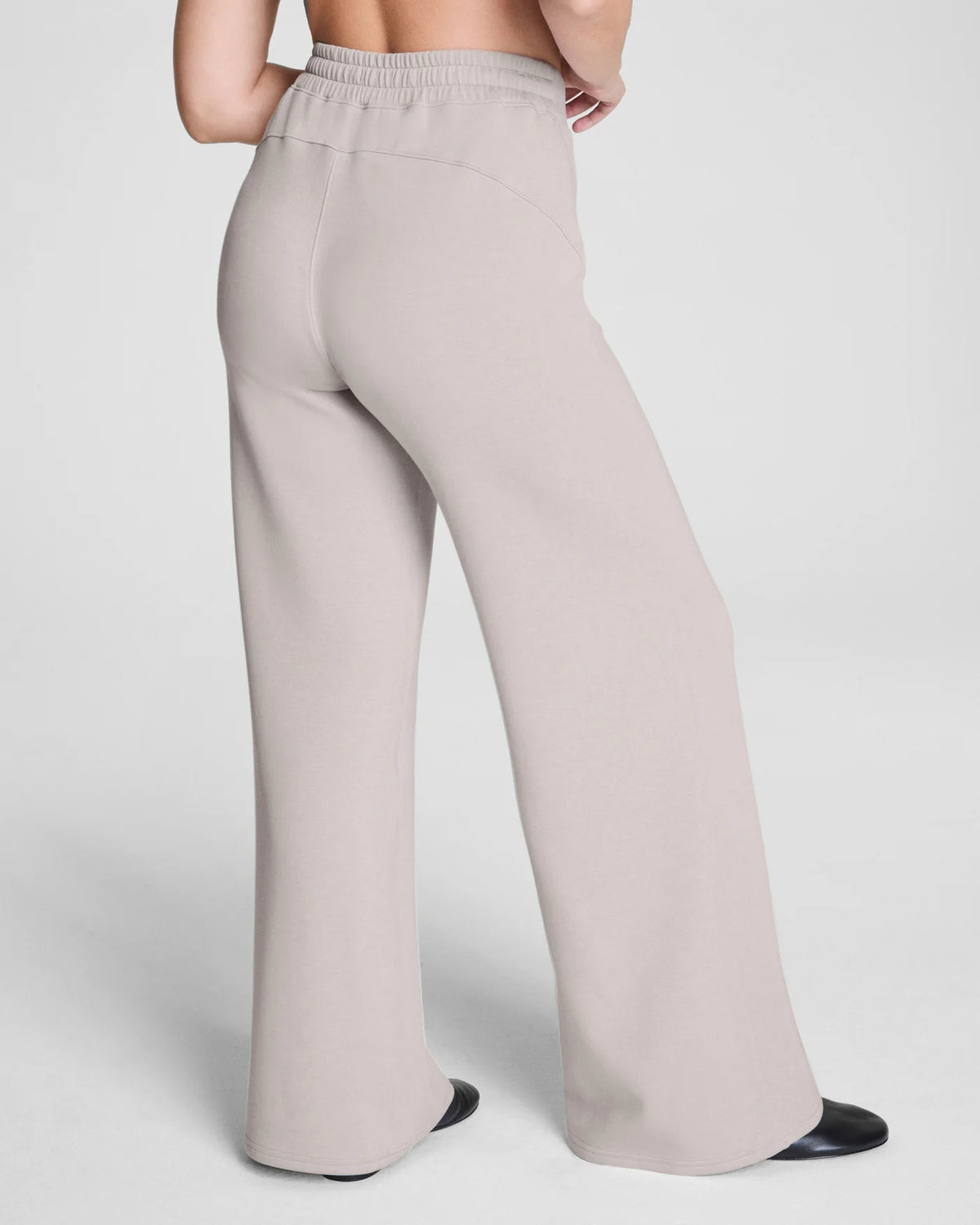 Someone stands facing away against a plain background, hands on hips, wearing SPANX - AIRESSENTIALS Wide Leg Pant in high-waisted, loose-fitting light beige and black shoes. The pants are crafted from airy fabric by SPANX.