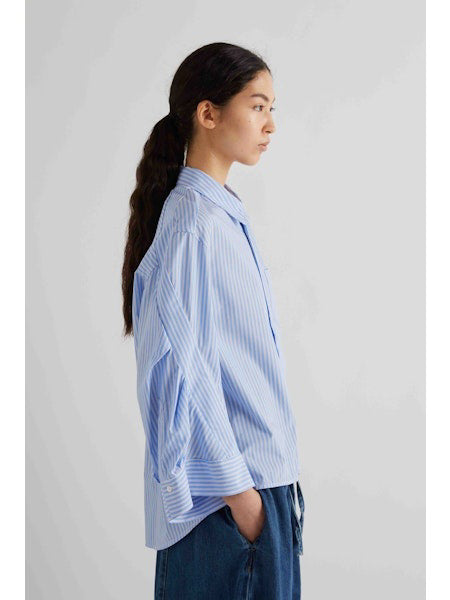 A woman with dark hair tied back models the MEIMEIJ STRIPE COTTON SHIRT—an oversized light blue and white striped cotton top with wide sleeves—paired with blue pants, standing against a plain light gray background.