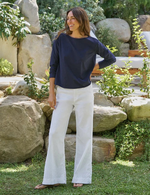 A woman stands outdoors, smiling with her hands in her pockets. She is wearing the FRANK & EILEEN Ella Long Sleeve Boatneck Tee in Classic Navy, paired with white pants. The background features rocks, greenery, and an outdoor lounge chair.