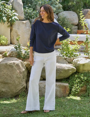 A woman stands outdoors, smiling with her hands in her pockets. She is wearing the FRANK & EILEEN Ella Long Sleeve Boatneck Tee in Classic Navy, paired with white pants. The background features rocks, greenery, and an outdoor lounge chair.