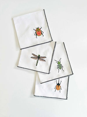 DOT AND ARMY’s EMBROIDERED INSECT COASTER set includes four linen/cotton coasters, each with a unique bug—red beetle, orange and black beetle, green dragonfly, and green bug—plus dark stitched edges, arranged on a white surface.
