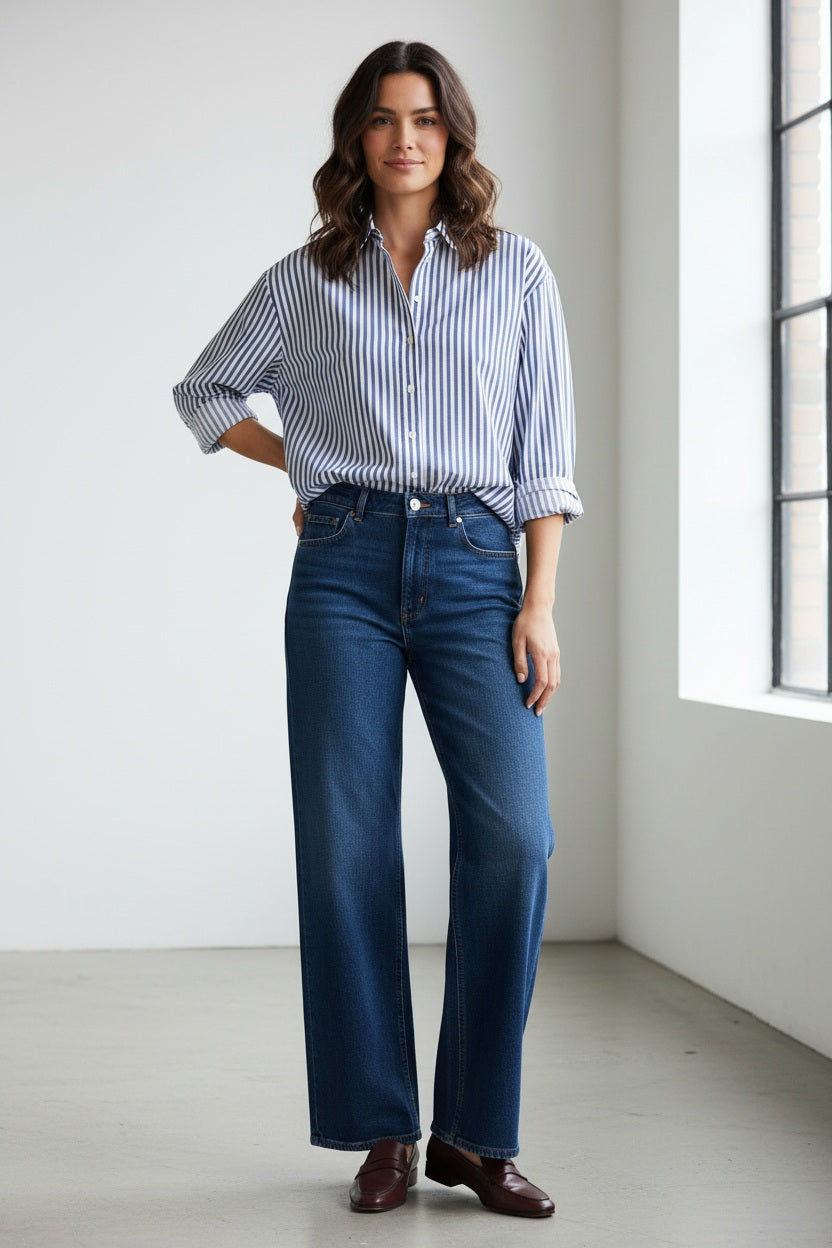 A woman with long brown hair stands against a plain background, smiling slightly. She is wearing a blue and white striped blouse paired with the SPANX Shape EveryWear Pull On Wide Leg Jean in Poseidon by SPANX and brown shoes, accessorized with a watch on her wrist.