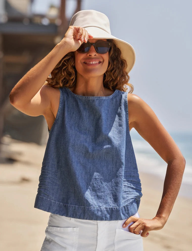 Wearing the FRANK & EILEEN SHELLEY Shell Tank in Famous Denim Distressed Indigo Wash, a woman stands barefoot on a sandy beach beside a large tote bag filled with white flowers, paired with white wide-leg pants, a sunhat, and sunglasses.