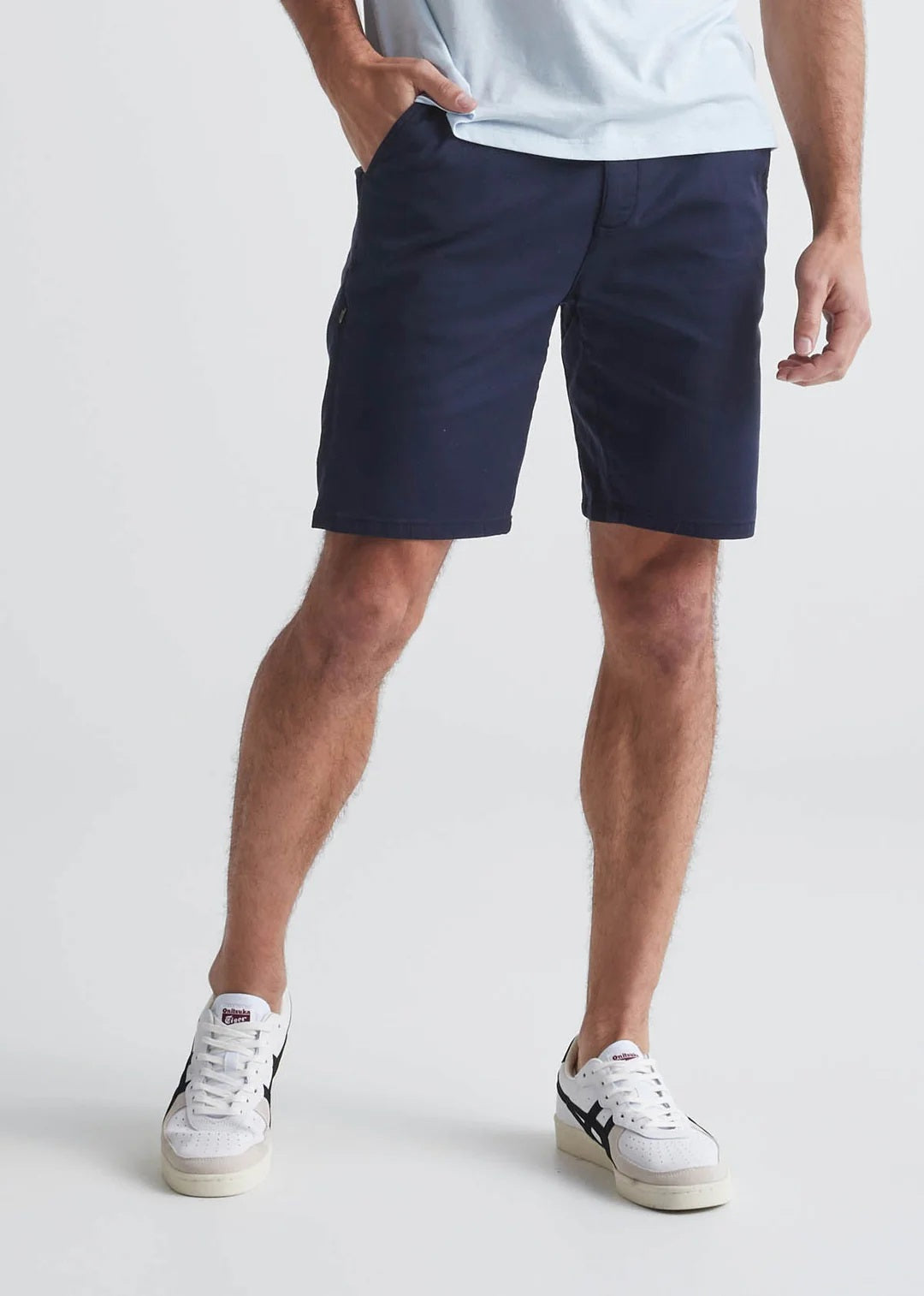 A person is walking against a plain light background, one hand in their pocket. Only the lower half is visible, showing gray t-shirt, white sneakers with black accents, and DUER's Live Free Journey Shorts in Sapphire.
