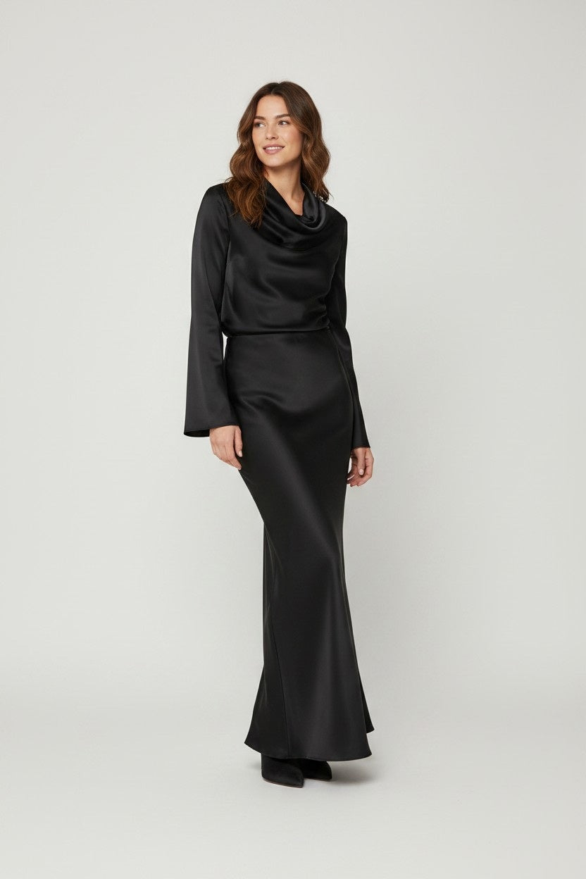 A woman stands smiling, wearing the MILVA MI - LONG SATIN SKIRT IN BLACK by MILVA, styled with a fluid silhouette. The skirt flows to the floor in lustrous black satin, paired with a fitted top that features long sleeves and a cowl neckline. She has wavy brown hair and wears black shoes, standing against a plain light background.