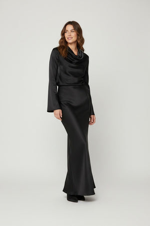 A woman stands smiling, wearing the MILVA MI - LONG SATIN SKIRT IN BLACK by MILVA, styled with a fluid silhouette. The skirt flows to the floor in lustrous black satin, paired with a fitted top that features long sleeves and a cowl neckline. She has wavy brown hair and wears black shoes, standing against a plain light background.