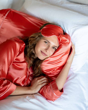 A woman in MERSEA’s Over The Moon Satin Sailor Sleep Mask sits on a bed, smiling gently as she lifts the mask from her eyes, with a softly blurred, lavender-toned background enhancing the calm mood.