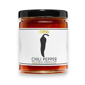 Two slices of bread topped with cream cheese and BRINS - CHILI PEPER SPREAD sit on a plate. Beside the plate is an open jar of BRINS Chili Peper Spread with a knife inside, all placed on a light surface with soft shadows.