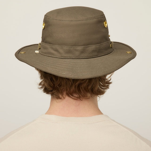 A young person with light skin and wavy brown hair wears the TILLEY - CLASSIC T3 HAT IN OLIVE and a beige crew neck shirt, looking seriously at the camera against a plain light background.