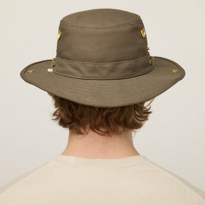 A young person with light skin and wavy brown hair wears the TILLEY - CLASSIC T3 HAT IN OLIVE and a beige crew neck shirt, looking seriously at the camera against a plain light background.