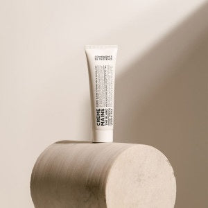 A bottle of Savon Liquide de Marseille soap, crafted with nourishing Provence plant oils, and a tube of COMPAGNIE DE PROVENCE - TRAVEL HAND CREAM WHITE TEA 1OZ by CIE LUXE BRANDS rest on a round tray atop a wooden stool, set against a backdrop of beige tiles.