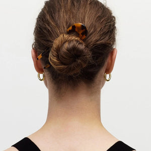 A person with brown hair in a low bun secured by the MACHETE - FRENCH HAIR PIN IN CLASSIC TORTOISE, wearing gold hoop earrings and a black top, is shown from the back against a plain white background.