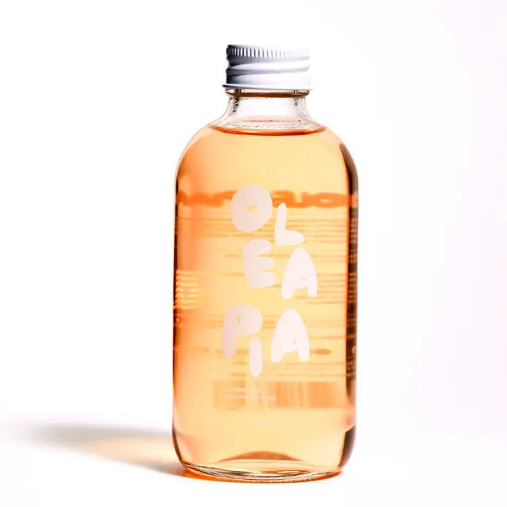 A clear glass bottle filled with light orange liquid, labeled "OLEA PIA - ROSE BALSAMIC VINEGAR" in white letters. It features a silver cap and is photographed against a simple white background. The design is minimalistic with a modern aesthetic, emphasizing its sweet and fruity vinegar essence from the brand OLEA PIA.