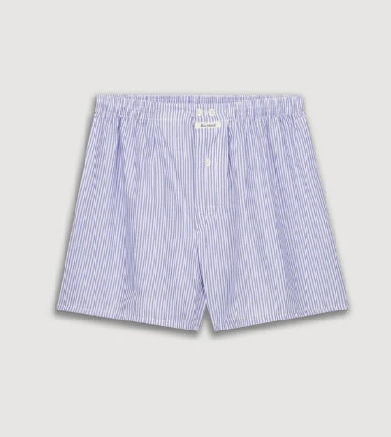 A person models HARTFORD - COTTON BOXER SHORTS in light blue and white stripes with an elastic waistband, standing against a plain beige background.
