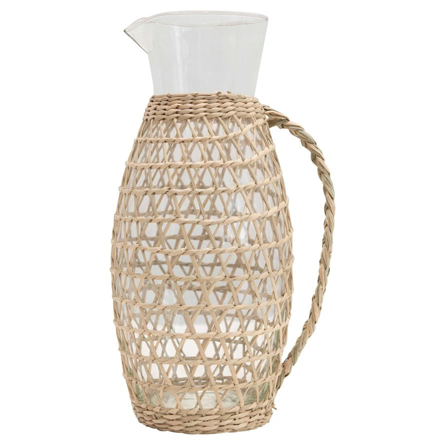 The CREATIVE COOP PITCHER WITH WOVEN SEAGRASS SLEEVE is a 2-quart clear glass pitcher featuring a handle and a spout that remains uncovered. It is elegantly encased in a seagrass weave cover extending from the base to the neck, fusing natural materials with modern functionality.