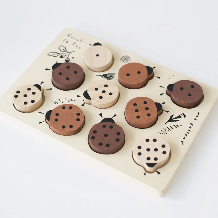 The WEE GALLERY - LADYBUGS COUNT TO 10 TRAY PUZZLE by WEE GALLERY is a wooden puzzle board featuring ladybug-shaped pieces in varying shades from light beige to dark brown. This educational puzzle helps teach counting skills and depicts leaves and twigs alongside the words "Count to Ten" on the left side. Each ladybug is adorned with black dots on their backs to represent number concepts.