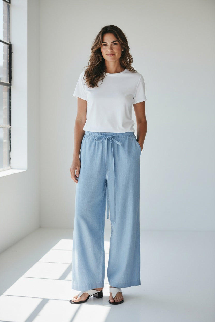 Model wears MEIMEIJ - DRAWSTRING COTTON PANTS in light blue, featuring a loose fit, wide legs, cropped ankle, and drawstring waist. Styled with white sandals. Image shows shoulders to feet against a plain background.