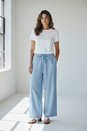 Model wears MEIMEIJ - DRAWSTRING COTTON PANTS in light blue, featuring a loose fit, wide legs, cropped ankle, and drawstring waist. Styled with white sandals. Image shows shoulders to feet against a plain background.
