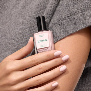 A hand with pale pink polished nails holds a bottle of MANUCURIST INC GREEN™ Nail Polish in Blossom against a gray knit sweater background.
