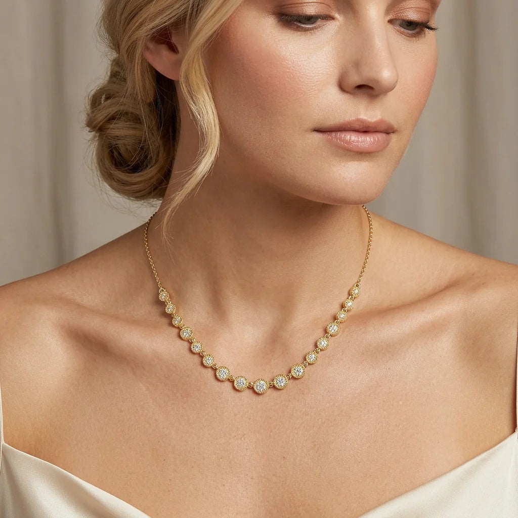 A woman with blonde hair styled in a low updo wears a white off-shoulder top and the ELLA STEIN - MULTI CIRCLE CHAIN NECKLACE by ELLA STEIN, featuring gold plated chains adorned with round, sparkling floral-shaped pendants. She looks slightly down with a calm expression against a neutral background.
