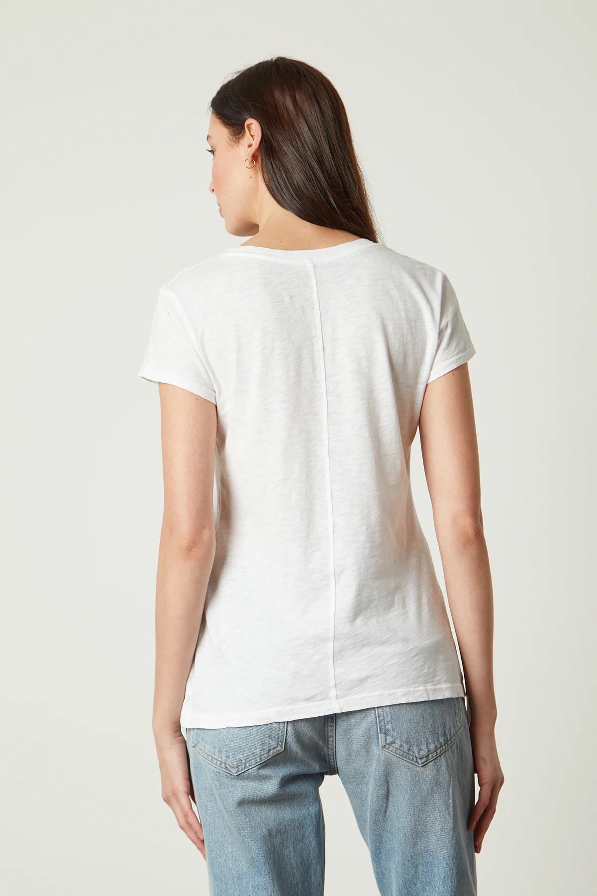 A woman with long brown hair stands sideways, looking towards the camera. She is wearing a hi-lo luxe essential beige VELVET - ODELIA CREW NECK COTTON SLUB TEE from VELVET BY GRAHAM &amp; SPENCER and dark blue jeans. Her left hand is in her pocket, and she stands against a plain white background.
