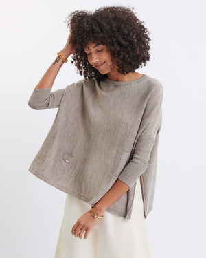 A woman with curly hair wears the MERSEA - CATALINA CREWNECK SWEATER IN DRIFTWOOD, a relaxed fit sweater by MERSEA, layered over a light skirt. She smiles gently while looking down, one hand touching her hair and the other resting on her wrist. The background is plain white.