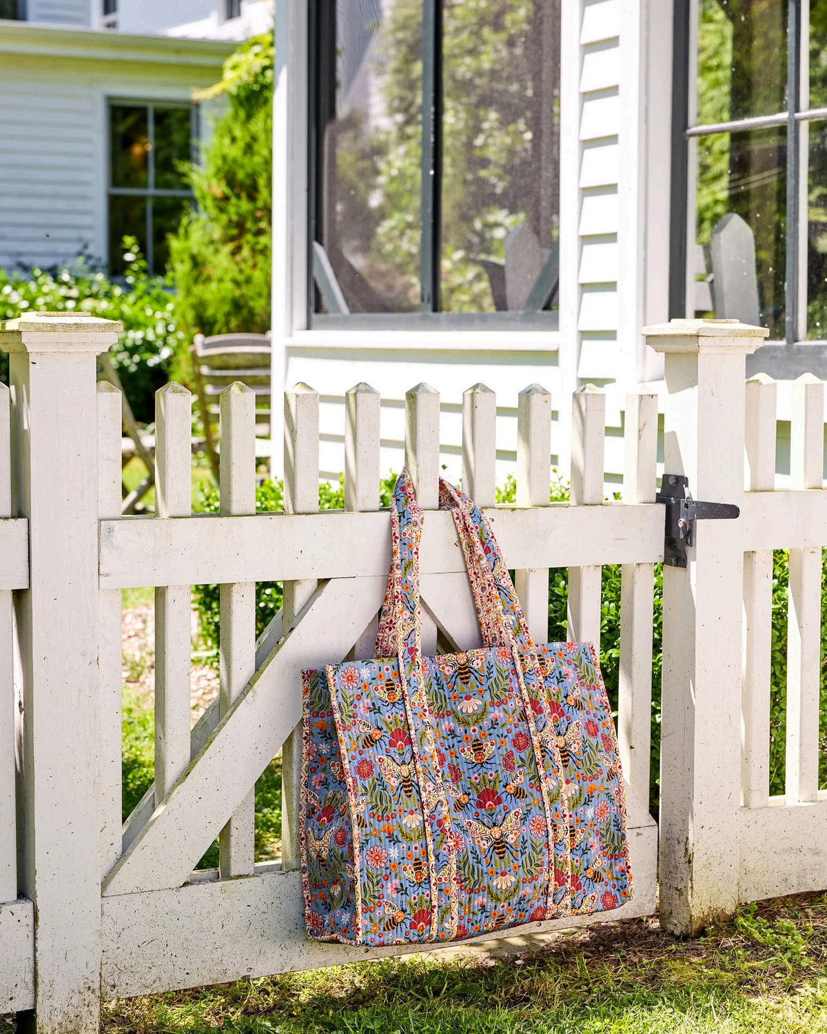 The PRINTFRESH - Bee Tapestry Traveler Tote in Delphinium Blue by PRINTFRESH, featuring vibrant floral, butterfly, and bee designs on organic cotton, hangs on a rustic wooden fence outdoors with lush green foliage in the background.