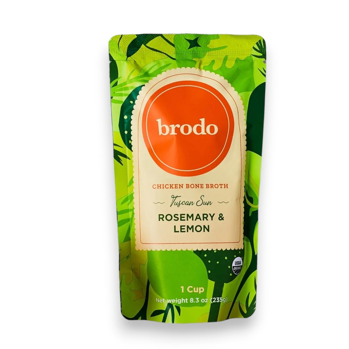 A pouch of BRODO BROTH COMPANY’s BRODO BROTH- TUSCAN SUN ROSEMARY & LEMON BONE BROTH ORGANIC sits on a white surface, surrounded by garlic, lemon, rosemary, and a bowl of broth. This keto-friendly and gluten-free bone broth delivers wholesome goodness to any kitchen.