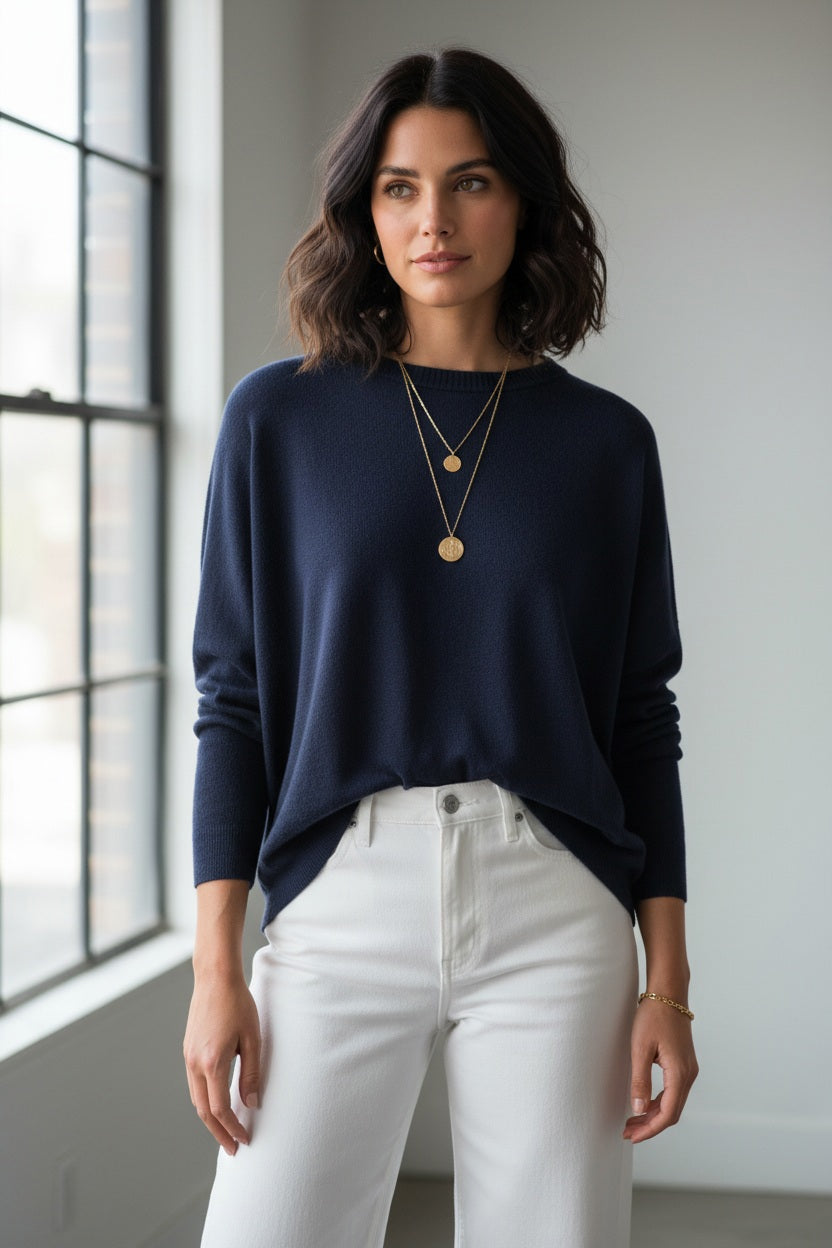 A woman stands in front of a gray wall and a green potted plant, wearing the MERSEA - CATALINA CREWNECK SWEATER IN NAVY by MERSEA with an oversized fit, paired with white pants, layered necklaces, and a bracelet, as she looks slightly to the side.