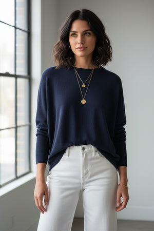 A woman stands in front of a gray wall and a green potted plant, wearing the MERSEA - CATALINA CREWNECK SWEATER IN NAVY by MERSEA with an oversized fit, paired with white pants, layered necklaces, and a bracelet, as she looks slightly to the side.