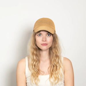A woman with long wavy blonde hair wears the WYETH HATS - PAULA MESH CAP IN CAMEL by WYETH, featuring UPF 50+ protection, and a white tank top, seated against a plain light-colored background.