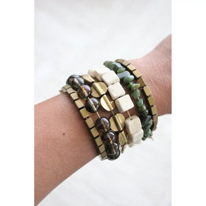 The DAVID AUBREY ATHENA HEMATITE GLASS AGATE JADE WRAP BRACELET, by DAVID AUBREY, features metallic gold square beads, transparent and ivory geometric accents—an elegant Mother's Day gift displayed on a gray background.