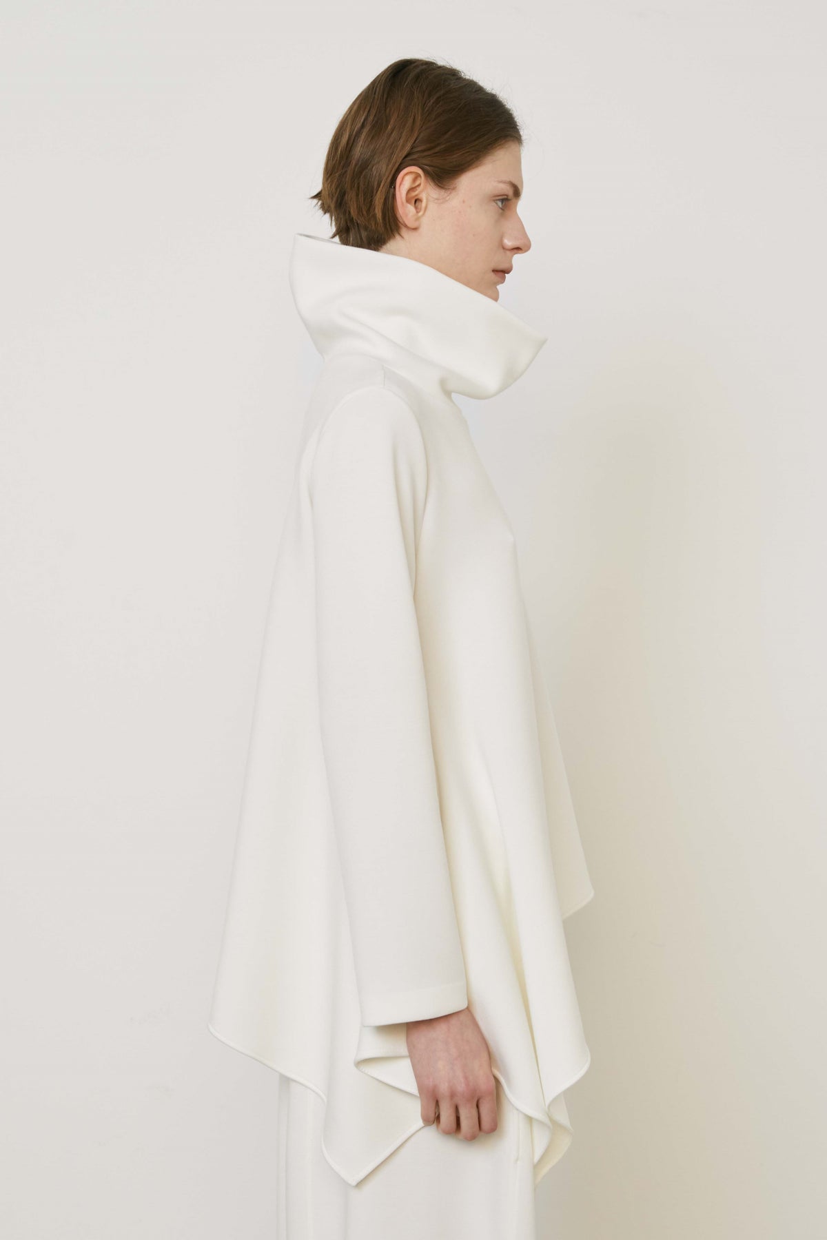 A person with brown hair faces away wearing the MEIMEIJ ASYMMETRICAL COWLNECK TOP in lightweight scuba fabric, featuring an asymmetrical hem, paired with matching white bottoms, against a plain white background.