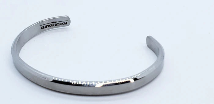 The CLIFTON WILSON - SILVER STAINLESS STEEL BANGLE is a minimalist, open-ended men's cuff featuring the brand engraved inside, shown against a white background for a refined accessory look.