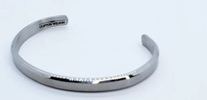 The CLIFTON WILSON - SILVER STAINLESS STEEL BANGLE is a minimalist, open-ended men's cuff featuring the brand engraved inside, shown against a white background for a refined accessory look.