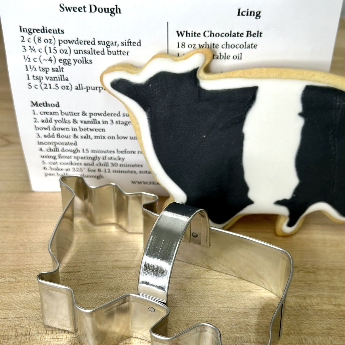 A cookie cutter from the FEARRINGTON SIGNATURE COLLECTION - BELTIE COOKIE CUTTER by AD RESOURCES is shaped like a cowboy boot, wrapped in clear plastic and tied with a black and white striped ribbon. Attached is a tag with the word &quot;FEARINGTON,&quot; an image of the cookie cutter, and a recipe card for Beltie Cow cookies.