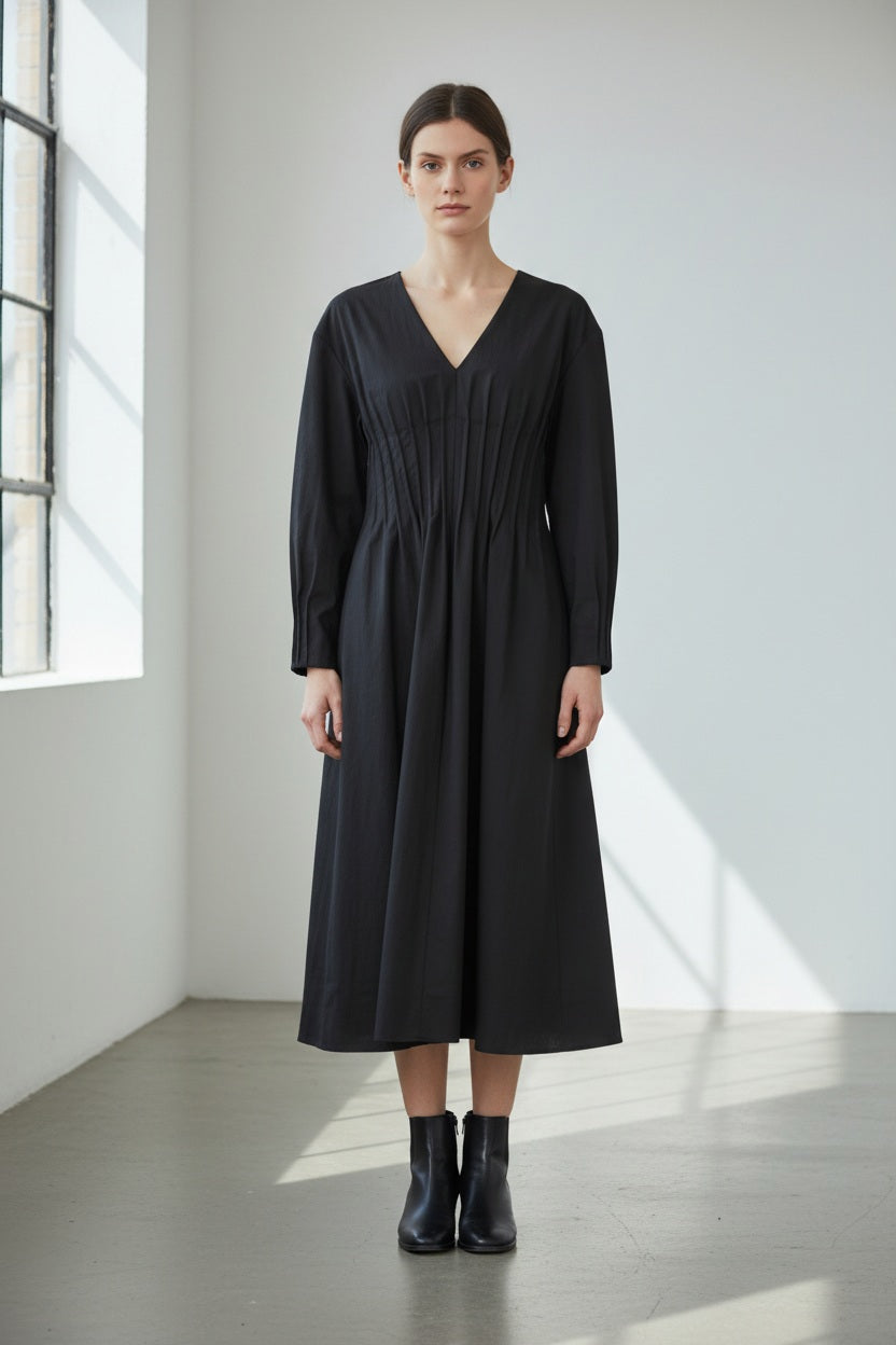 A person stands indoors wearing the MEIMEIJ Gathered Waist Dress in black—a long-sleeved, V-neck pleated midi dress crafted in Italy—paired with black ankle boots. The minimal space features white walls and large windows that fill the room with natural light.