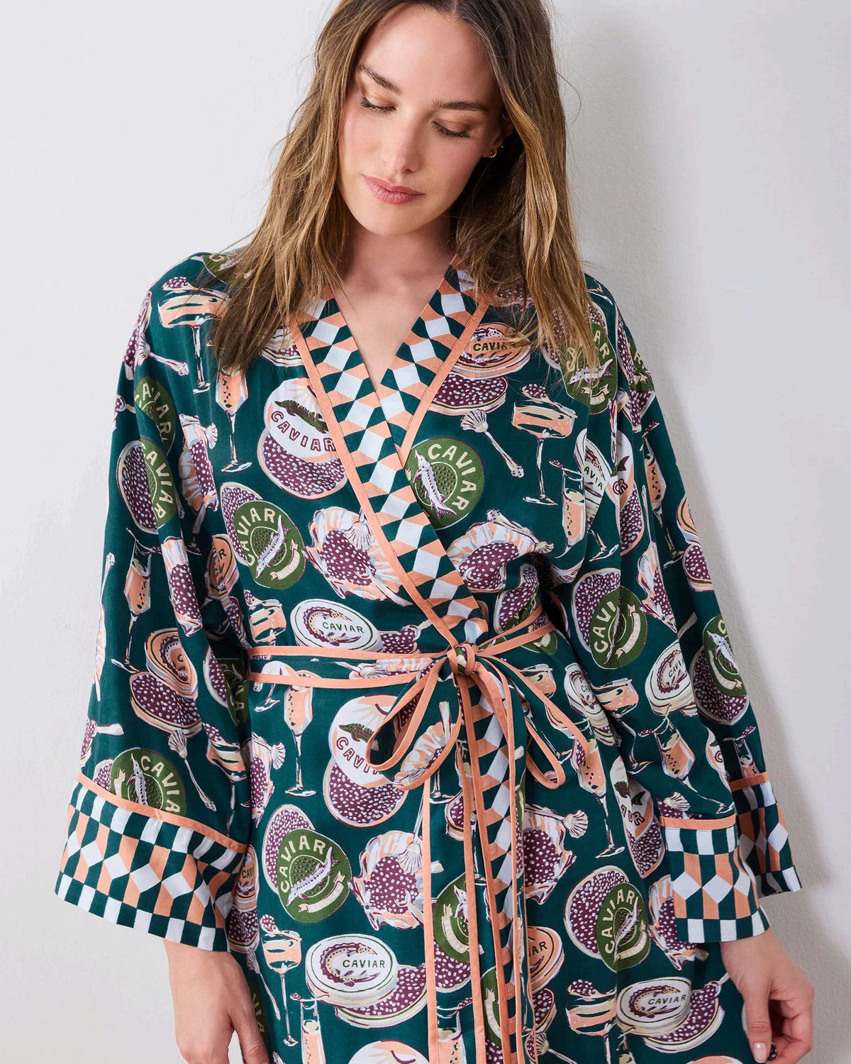 A woman stands barefoot against a white wall, smiling and holding a white cup in one hand as she poses slightly angled to the side. She is wearing the PRINTFRESH Champagne & Caviar Eco Satin Robe in Peacock Feather—a long, colorful eco-friendly robe featuring a vibrant botanical print with peacock feathers and checkered trim.