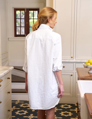 The FRANK AND EILEEN - MARY SHIRTDRESS IN TATTERED DENIM from FRANK & EILEEN is showcased against a textured, off-white background. This long-sleeved dress boasts a collared neckline, a single chest pocket, and a slightly curved hemline. The sleeves are rolled up neatly, making it an ideal favorite for all four seasons.